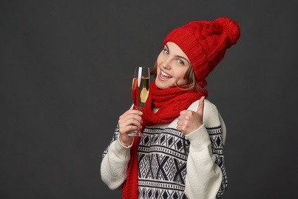 Winter holidays celebration. Portrait of young female in warm winter clothing holding a glass of champagne gesturing thumb up, over grey background